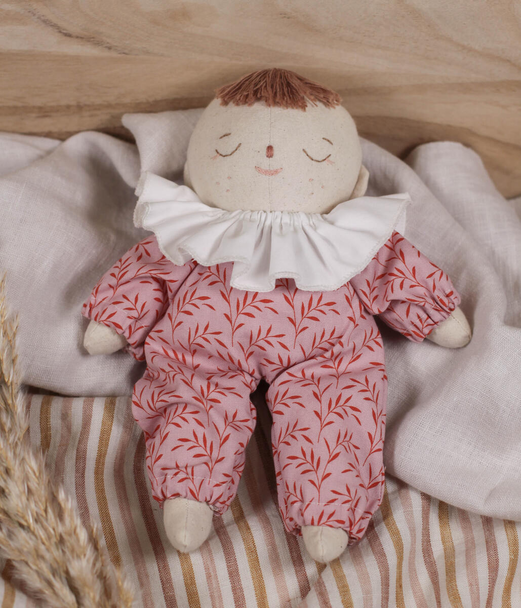Little Lenni baby doll Emily, sleeping on linen blanket dressed in floral pink overalls and with white ruffle collar around her neck.