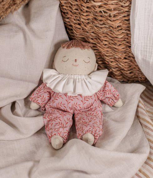 Little Lenni baby doll Emily, sleeping on linen blanket dressed in floral pink overalls and with white ruffle collar around her neck.