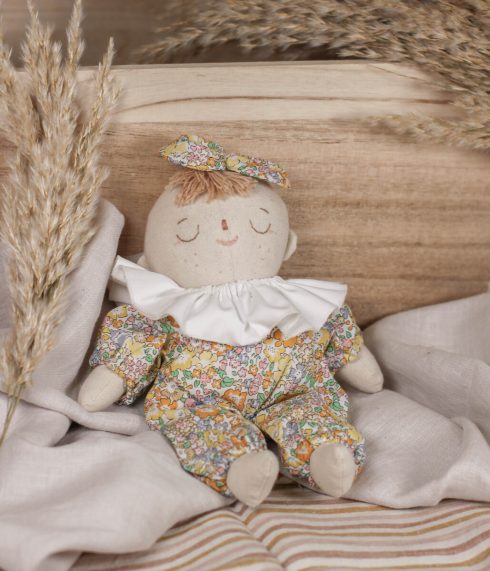 Little Lenni baby doll Lucy, sleeping in front of a basket dressed in floral yellow overalls and with white ruffle collar around her neck.