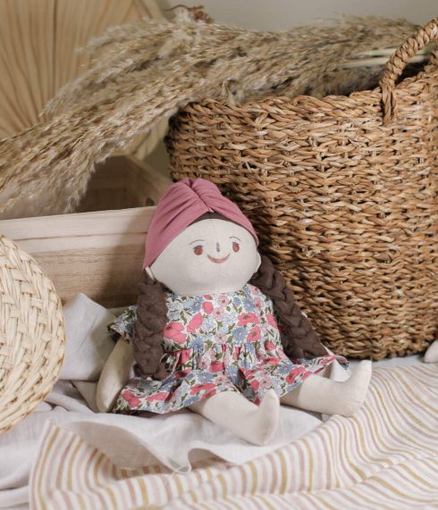 Little Lenni lutka Mila, sa pletenom tamnosmeđom kosom i očima boje lješnika, sjedi u cvjetnoj haljinici i šarenom turbanu. Ručno rađena od prirodnih i organskih materijala.