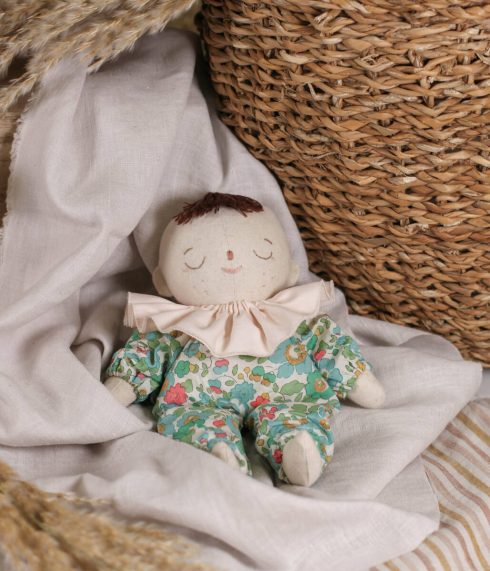 Little Lenni baby doll Pauline, sleeping in front of a basket dressed in floral green overalls and with light pink ruffle collar around her neck.
