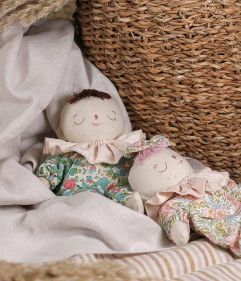 Two Little Lenni baby dolls, Pauline and Rosie, sleeping dressed in floral green and pink overalls and with pastel pink ruffle collars around their necks.