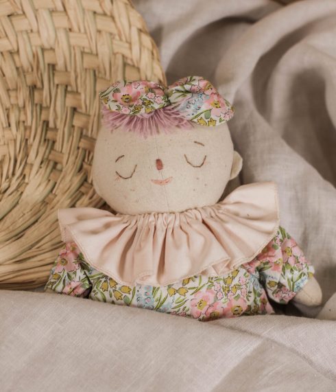 Little Lenni baby doll Rosie, sleeping covered with a blanket, and dressed in floral overalls and with pastel pink ruffle collar around her neck.